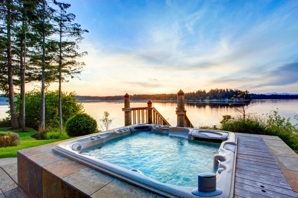 A hot tub on a patio overlooks a calm lake at sunset, surrounded by trees and greenery.