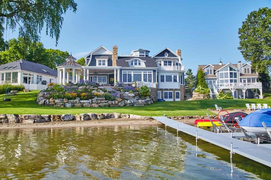 Large upscale waterfront home with gray-blue siding and white trim overlooking a calm lake. The property features a tiered stone retaining wall with landscaped flower beds, a covered patio, expansive lawn, private dock, and neighboring lakeside homes under a clear blue sky.